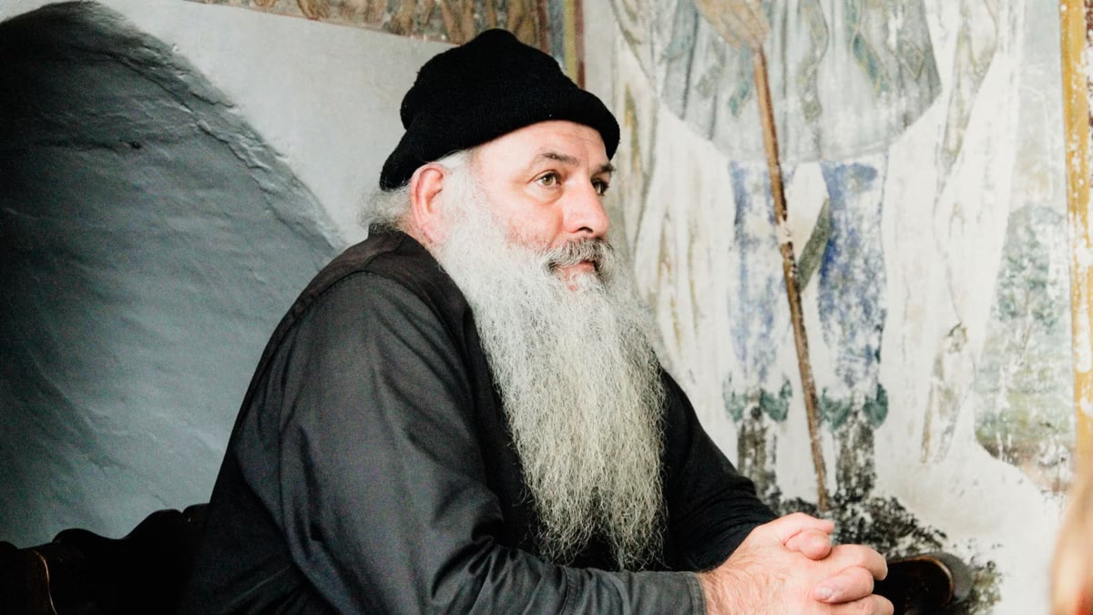 Elderly man with a beard wearing a beanie, seated inside a Greek monastery.