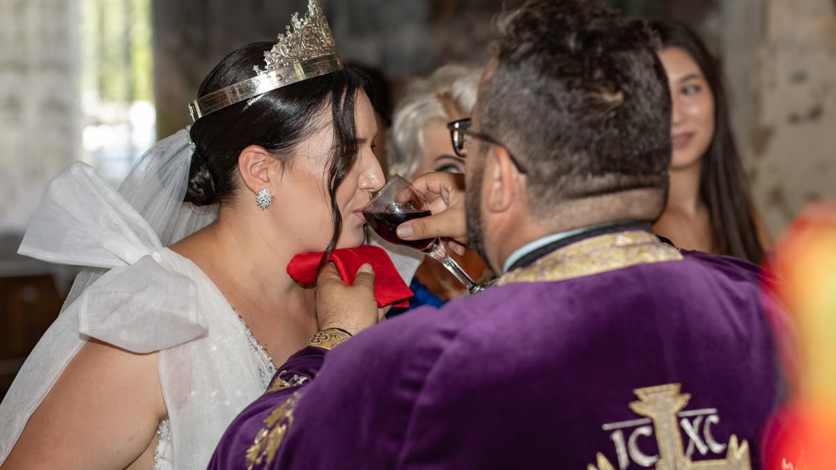 Bride wearing crown receives communion from priest during wedding ceremony indoors.
